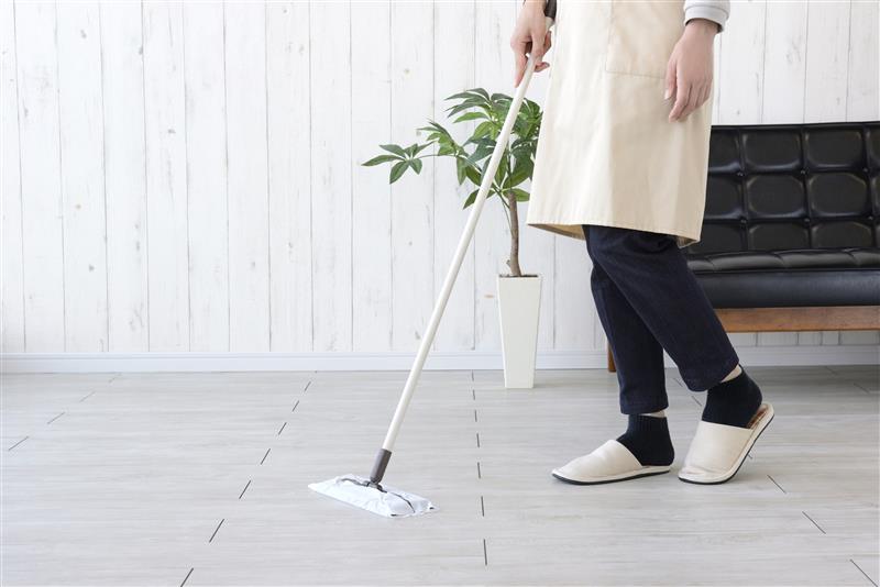 A waist-down shot of a person in an apron and slippers cleaning an already pristine floor with a flat mop. The image, set in a tidy, minimalist room, illustrates the article's point about Japanese participants meticulously cleaning their homes in preparation for a researcher's visit.