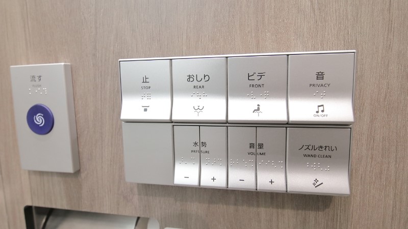 Control panel of a Japanese high-tech toilet (Washlet) featuring buttons for Stop, Rear, Front, Privacy Sound, Water Pressure, Volume, and Wand Clean, with Braille labels included.