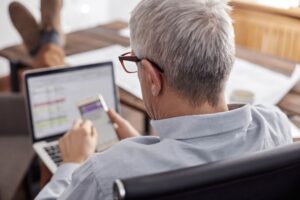 ノートパソコンとスマートフォンを操作するシニア男性の後ろ姿。複数のデバイスを使いながら業務に取り組む様子は、アクセシビリティとデジタルインクルージョンの重要性を象徴している。