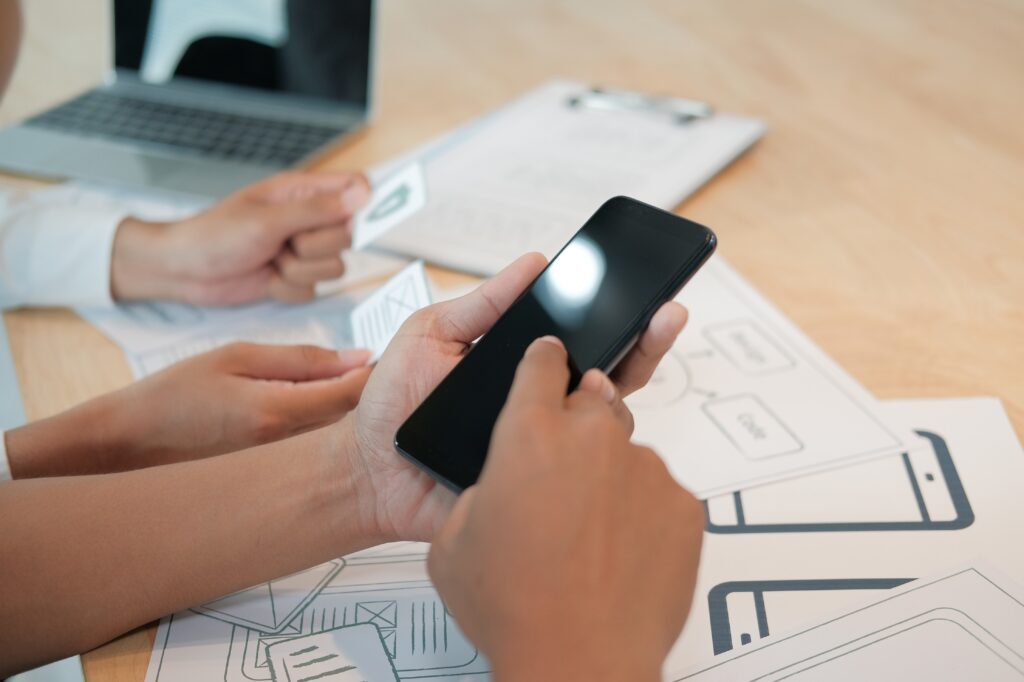 A close-up of a user participating in a usability test, holding a smartphone and interacting with its screen. Wireframe sketches and notes are visible on the desk, representing the process of product testing and user research.