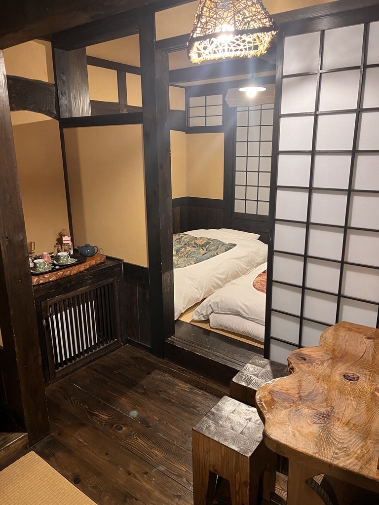 A traditional Japanese-style room in a ryokan. In the back is a sleeping area with futons laid out, and in the foreground are a shelf with a tea set and a wooden table and chairs.