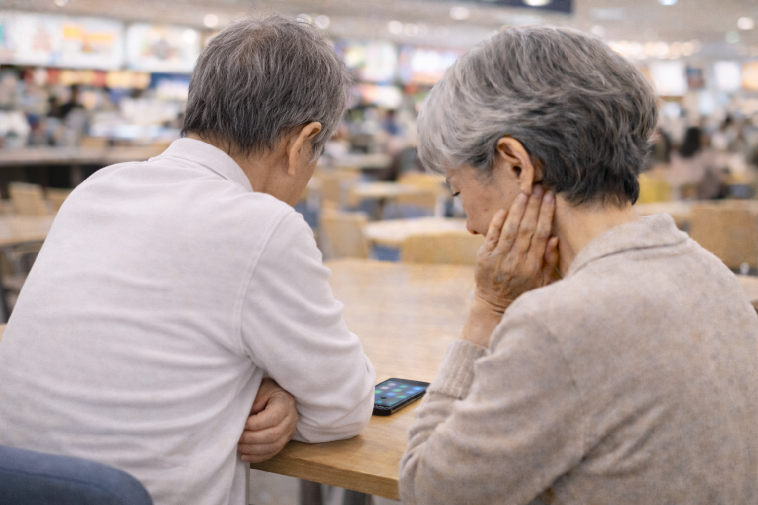 フードコートで一台のスマートフォンを前に困惑する高齢者夫婦の後ろ姿。記事冒頭で語られる、デジタルサービスの使い方に戸惑い、社会の利便性から取り残されがちな人々の課題を象徴している。