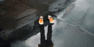 A person’s shadow is cast on wet pavement, with only a pair of orange shoes visible, symbolizing the article's theme of disappearing design.