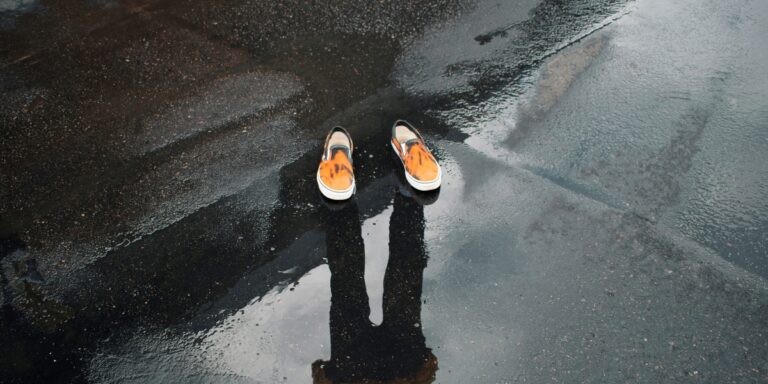A person’s shadow is cast on wet pavement, with only a pair of orange shoes visible, symbolizing the article's theme of disappearing design.