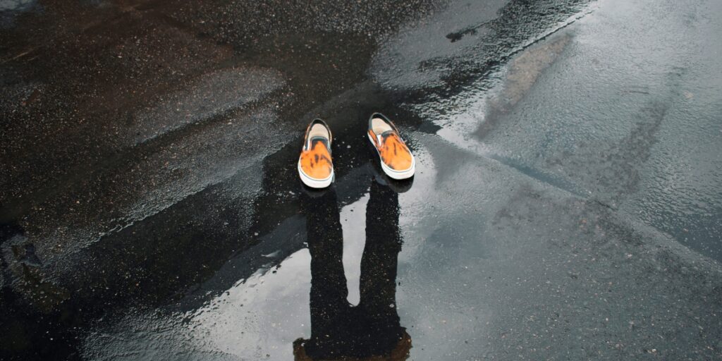 A person’s shadow is cast on wet pavement, with only a pair of orange shoes visible, symbolizing the article's theme of disappearing design.