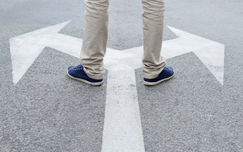 A person stands at a fork in the road marked by two large arrows, symbolizing the critical need for clear decision-making and strategic direction in the age of AI.