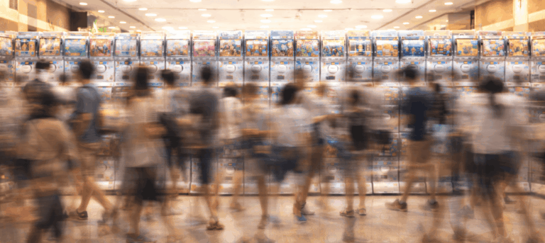 A long-exposure photograph showing a bustling crowd of people as motion blurs in front of a long, brightly lit wall of Japanese gashapon (capsule toy) machines.