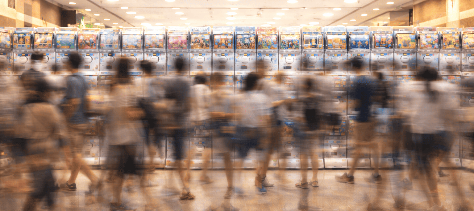 A long-exposure photograph showing a bustling crowd of people as motion blurs in front of a long, brightly lit wall of Japanese gashapon (capsule toy) machines.