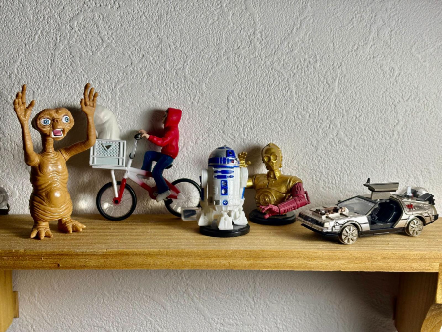 A collection of pop culture gacha toy figurines displayed on a wooden shelf against a white wall. From left to right, the figures are E.T., Elliott on his bicycle, R2-D2, a bust of C-3PO, and the DeLorean time machine from "Back to the Future."