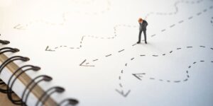 A miniature businessman stands on a notebook with multiple confusing, hand-drawn paths, representing the strategic challenge of choosing the right direction when AI generates endless options.