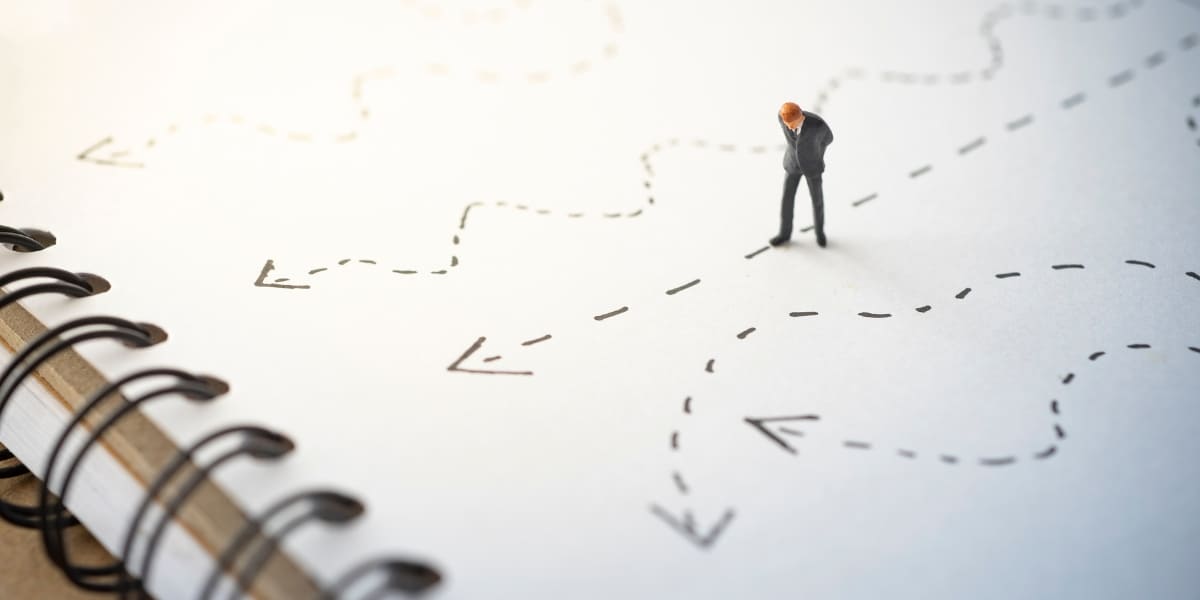 A miniature businessman stands on a notebook with multiple confusing, hand-drawn paths, representing the strategic challenge of choosing the right direction when AI generates endless options.