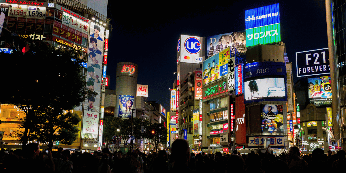 A photo of Tokyo's crowded Shibuya Crossing at night, its buildings covered in bright neon signs, representing the bustling social landscape that Japan's Gen Z is navigating on their own terms.