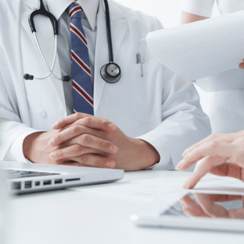 白衣を着た医師が、机を挟んでHFE評価の説明を受けている。腕を組んで話を聞く姿は、文化的背景によってフィードバックの出方が異なることや、日本の医療従事者への調査における丁寧なモデレーションの重要性を示唆している。