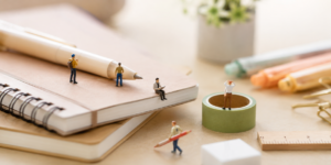A close-up, soft-focus shot of stationery items on a desk, including notebooks, a pen, and washi tape. Miniature human figures are placed around the items, interacting with them as if in a tiny world, which visually represents the theme of personal curation.
