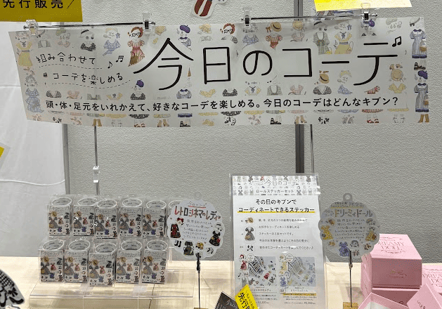 A booth display at the stationery expo centered around the theme '今日のコーデ' (Today's Outfit). A poster with this title is surrounded by products like mix-and-match stickers featuring illustrated cats in various outfits, promoting the idea of curating stationery.