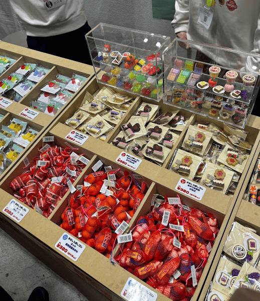 A display at a stationery expo booth filled with novelty erasers designed to look like food. There are bins of erasers packaged in mesh bags like fruit, and others in display cases shaped like Japanese sweets and sushi.