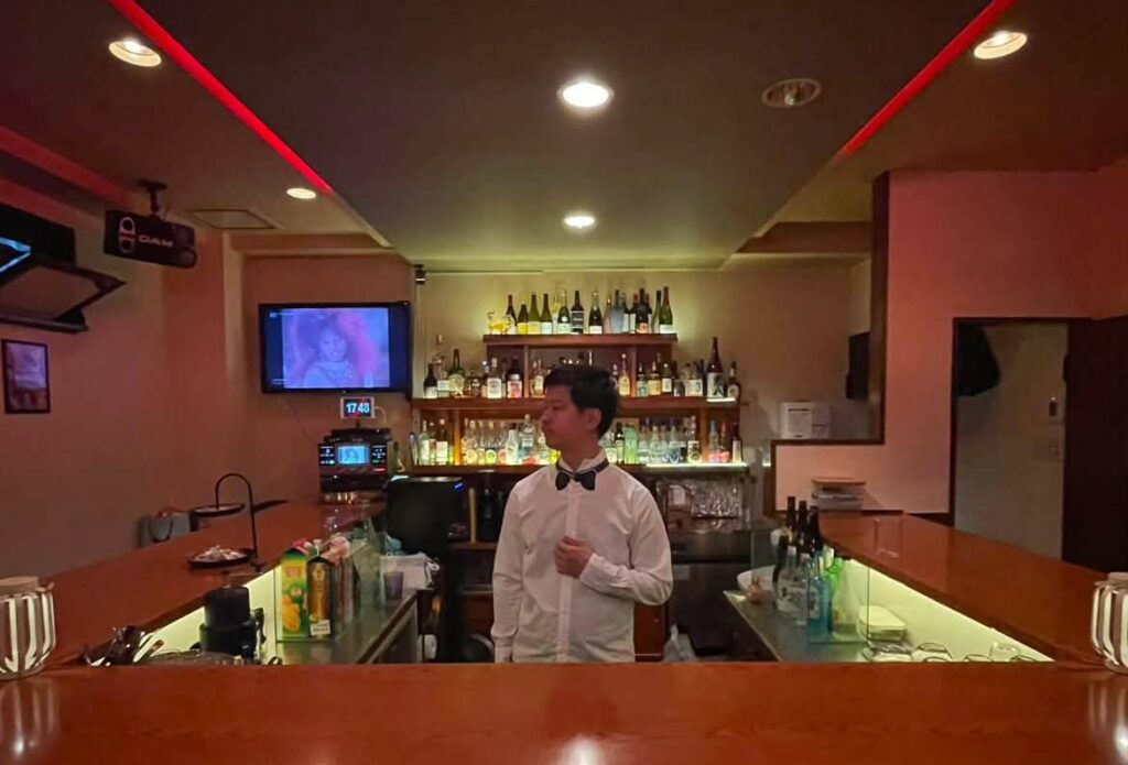 The brightly lit interior of the bar with a bartender standing in front of an easily visible selection of alcohol.