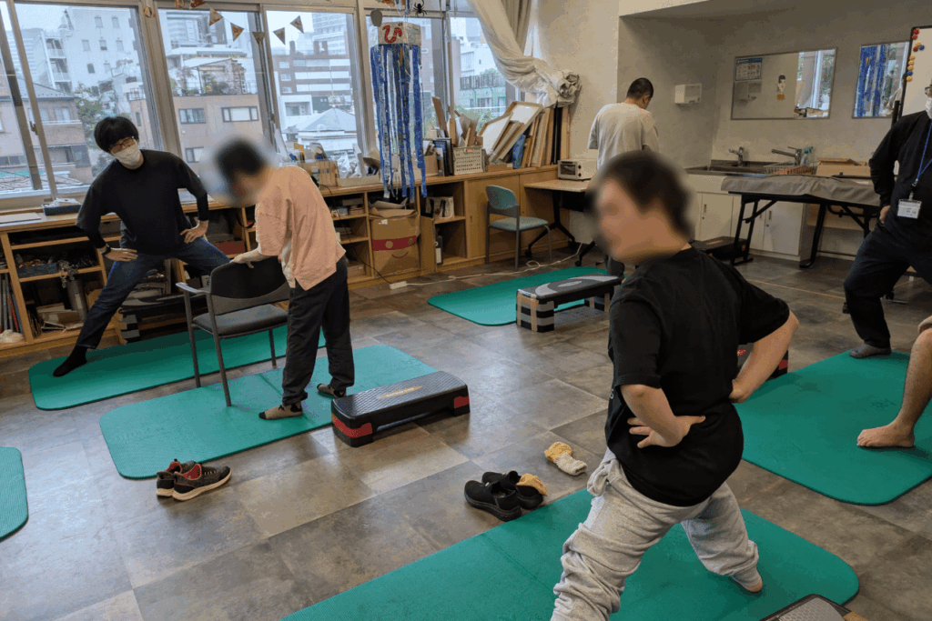 くるるえびすの室内で、マットの上に立った参加者たちがスタッフと一緒にストレッチ運動をしている様子。
